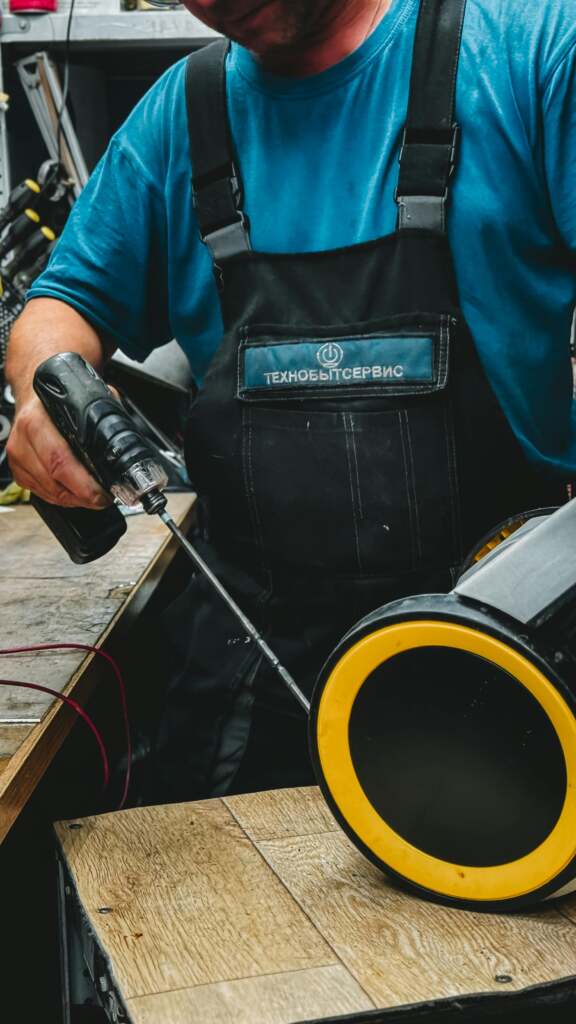 Technician in overalls using a drill for industrial machine repair in workshop.